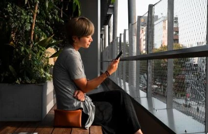 Mulher jovem sentada em café com vista para a rua, usando celular e refletindo, ideal para quem busca momentos de distração e conexão digital.