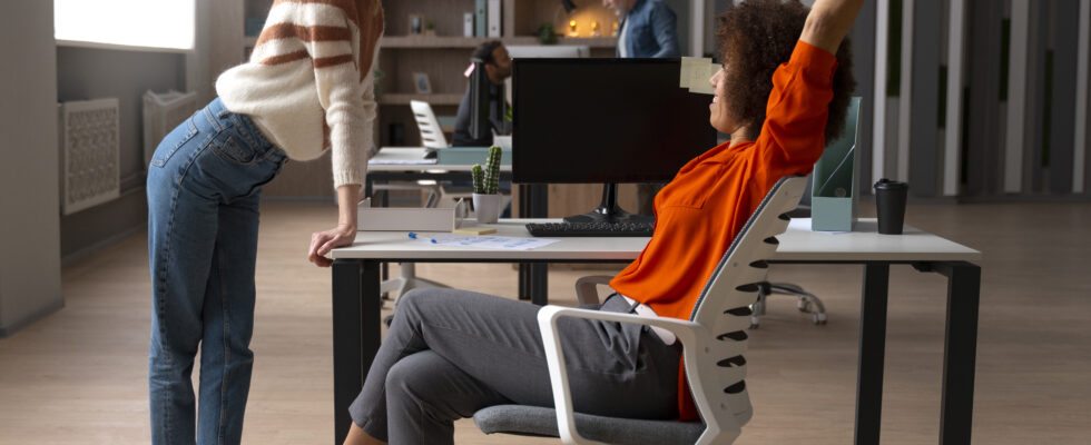 Duas mulheres interagindo de forma profissional em um escritório moderno, uma de pé e outra sentada na cadeira giratória, com expressão de conversa amigável.